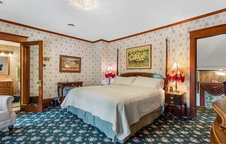 A floral patterned bedroom with a four-poster bed, two lamps, and an open doorway revealing a bathroom vanity.