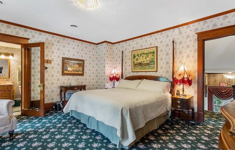 A floral patterned bedroom with a four-poster bed, two lamps, and an open doorway revealing a bathroom vanity.