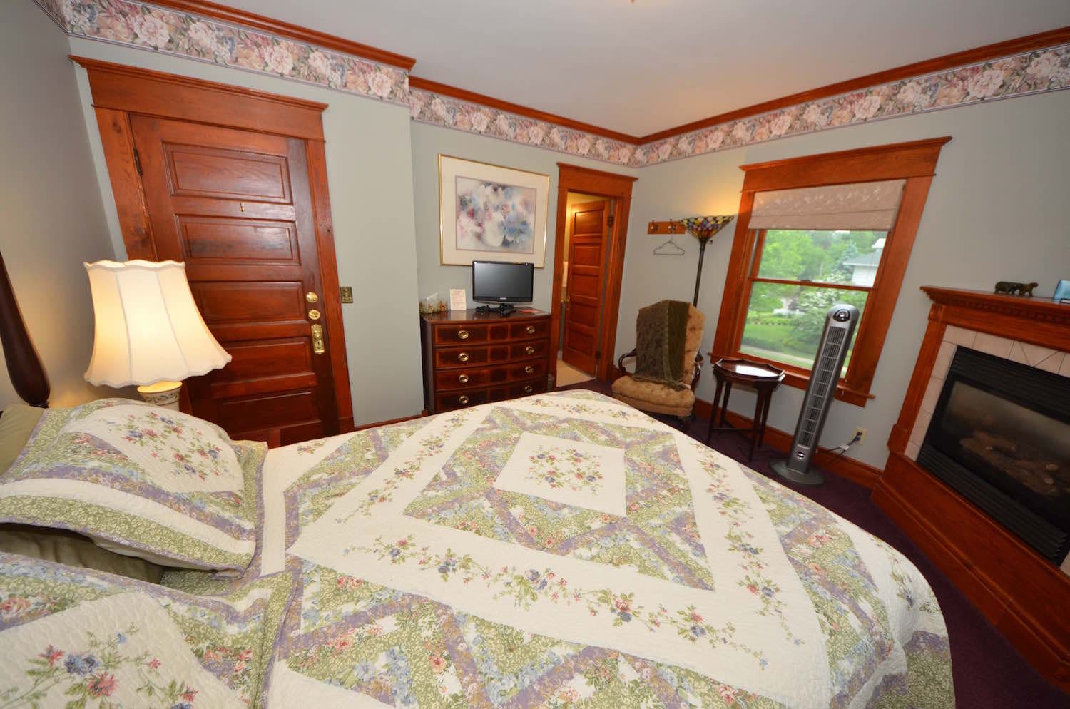 Bedroom with a large bed with a floral comforter, a fireplace, and a light purple wall with artwork.