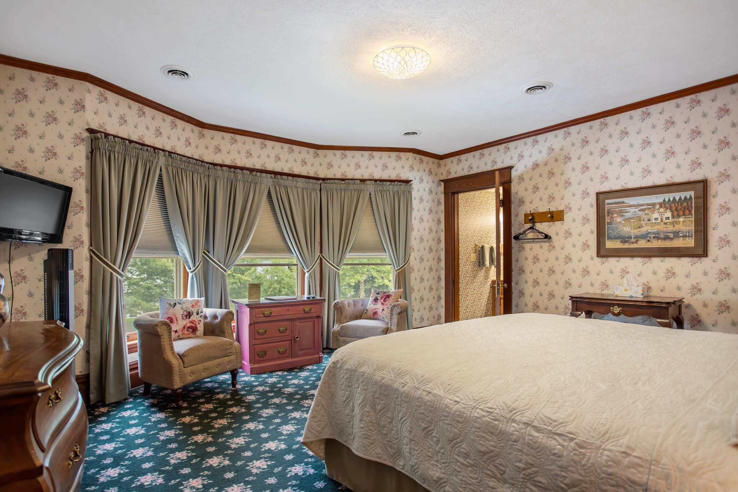 A bedroom with floral wallpaper, a large bed, two armchairs by a window, and a television.