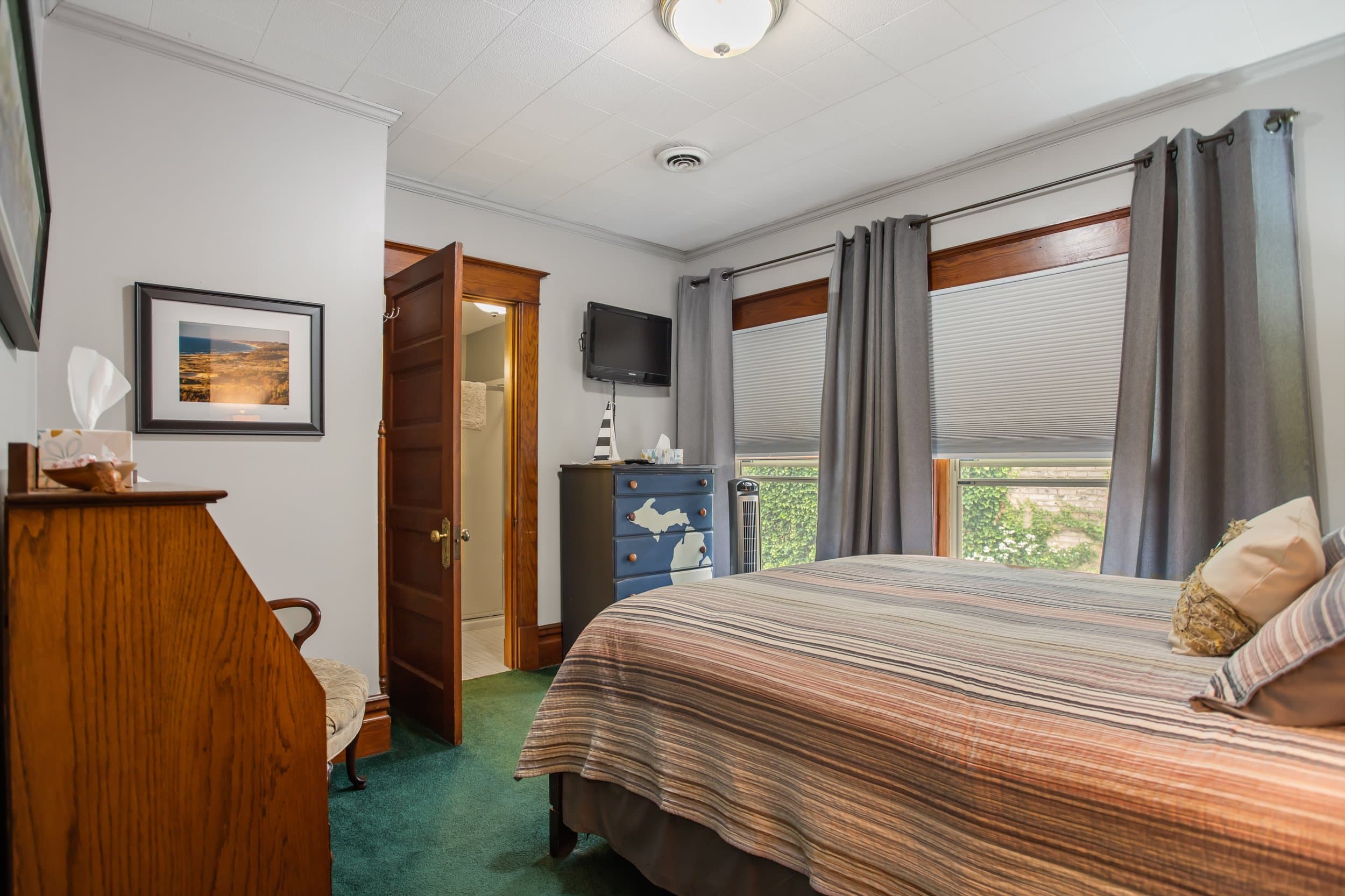 Bedroom with a striped comforter, a television, a dresser, and two windows with gray curtains.