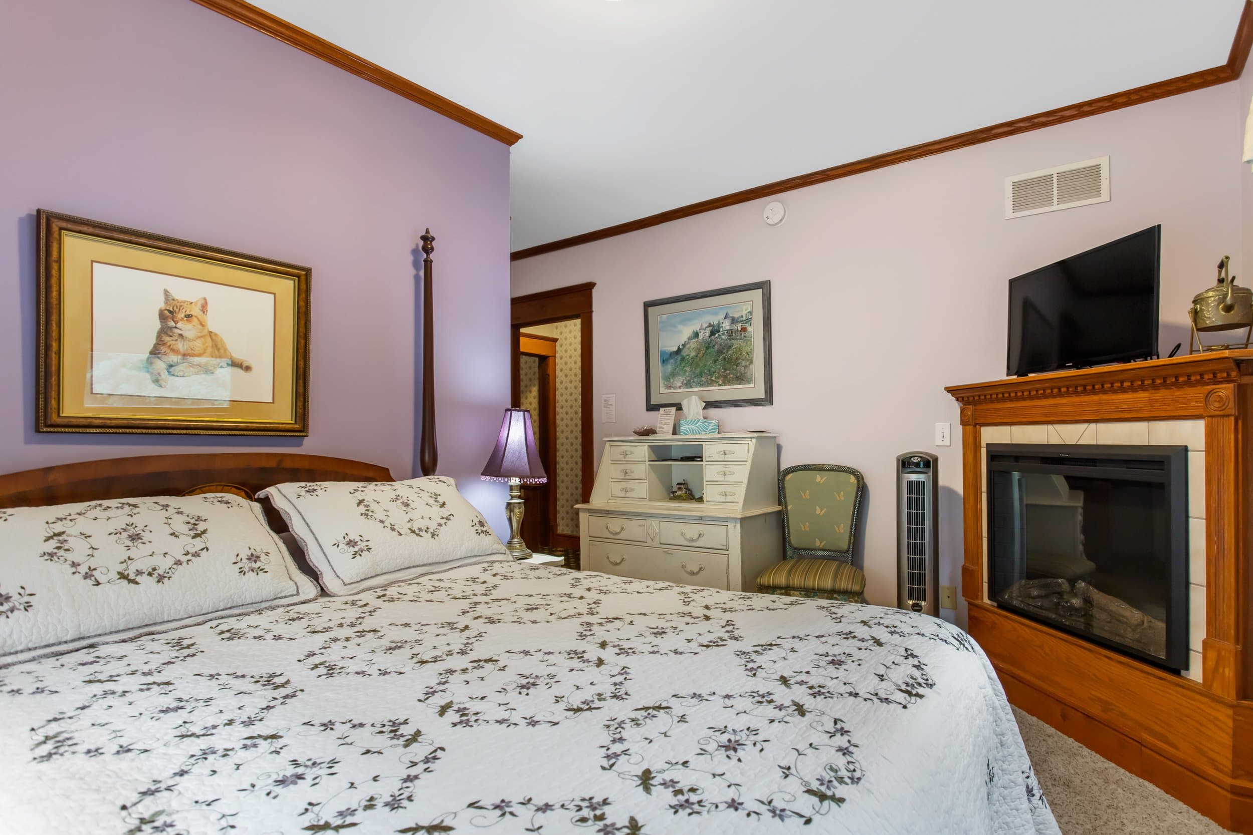 A cozy bedroom with soft lavender walls, a queen bed with a white patterned quilt, a wooden headboard, a fireplace, and a flat-screen TV.