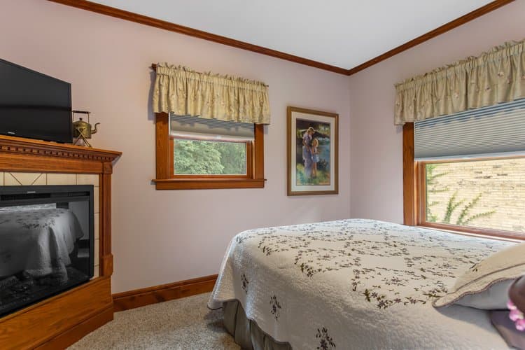 A soft lavender bedroom featuring a bed with a white patterned quilt, two windows with valances, a wooden fireplace with a TV above it, and a framed picture on the wall.