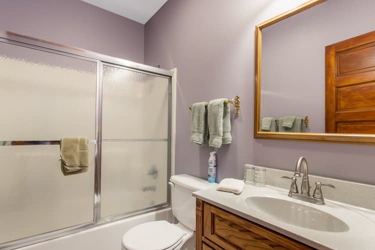 A bathroom with soft lavender walls, a shower-bathtub combo, a toilet, a wooden vanity with a sink and a large mirror, and towels hanging on a rack.