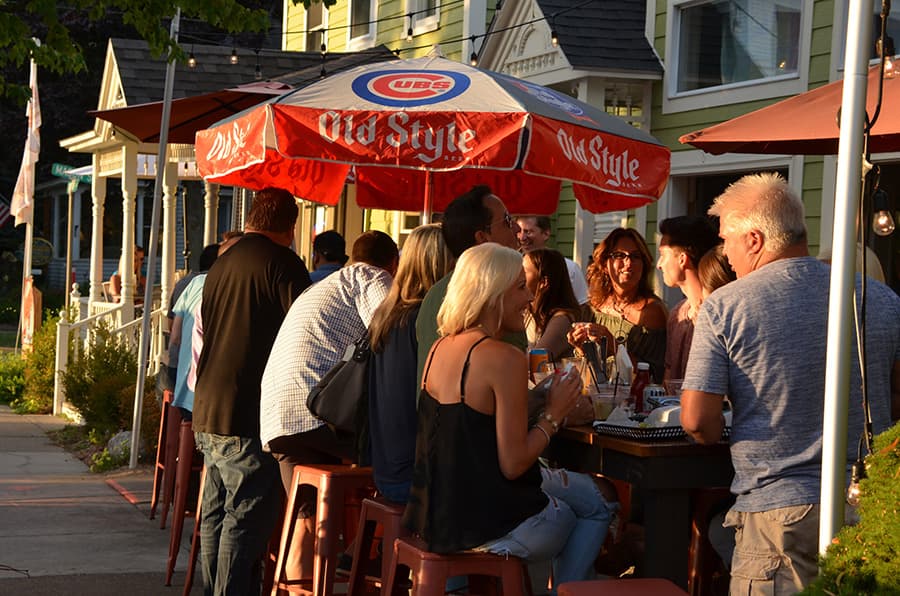 A group of people socializes at an outdoor bar under a colorful umbrella.