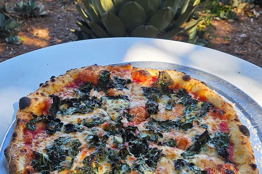A close-up of a pizza topped with leafy greens, sitting on a white table outdoors.