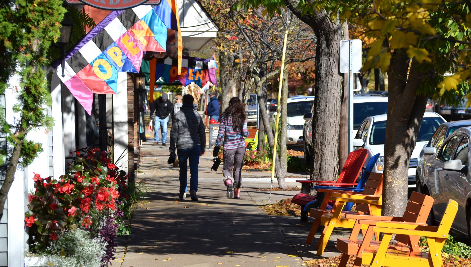 A bustling sidewalk adorned with colorful "OPEN" banners, potted flowers, and people walking by.