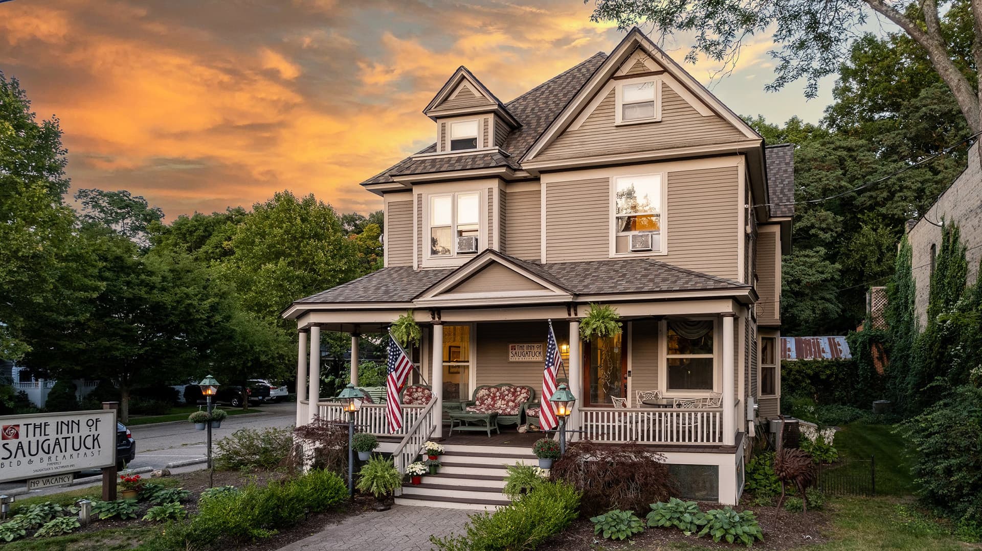 A charming bed and breakfast with a wraparound porch, flanked by trees, under a dramatic sunset sky.