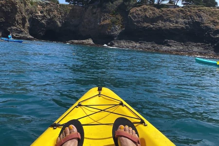A pair of feet in sandals resting on the bow of a yellow kayak in calm blue water, with a rocky shoreline and another kayaker in the background.